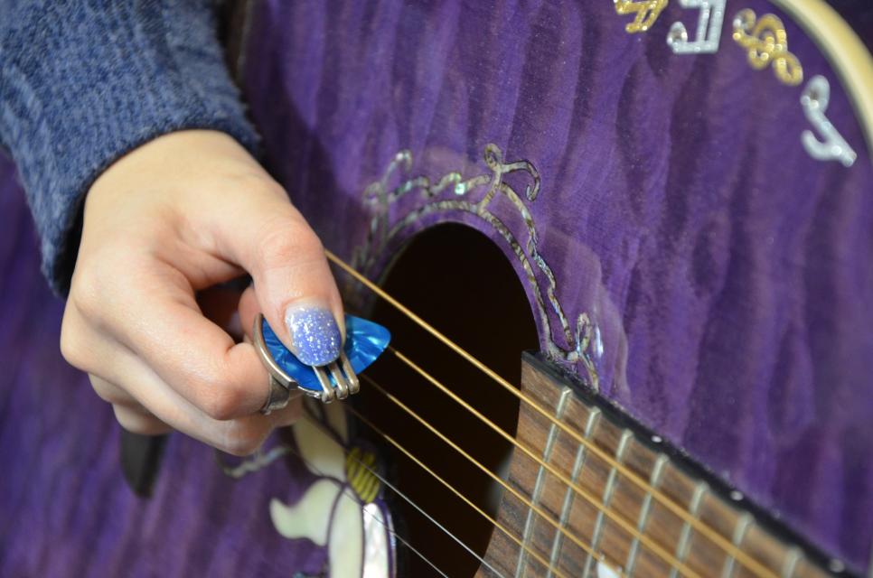 An image of a hand holding a guitar pick against the strings of a purple guitar. 