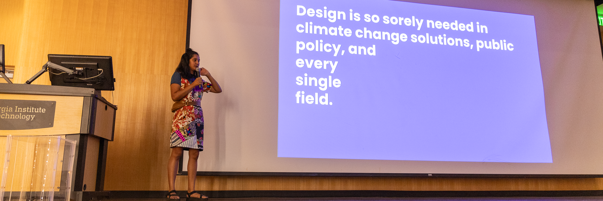 Krystal Persaud on stage at Reinsch Pierce Auditorium. A projection screen beside her reads, "Design is so sorely needed in climate change solutions, public policy, and every single field."