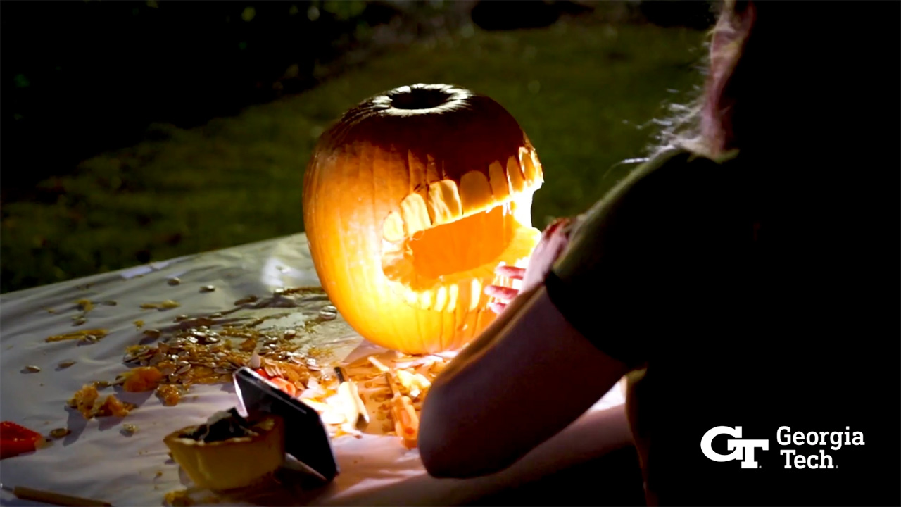 Video still of a carved pumpkin lit by a smartphone light while a student carves