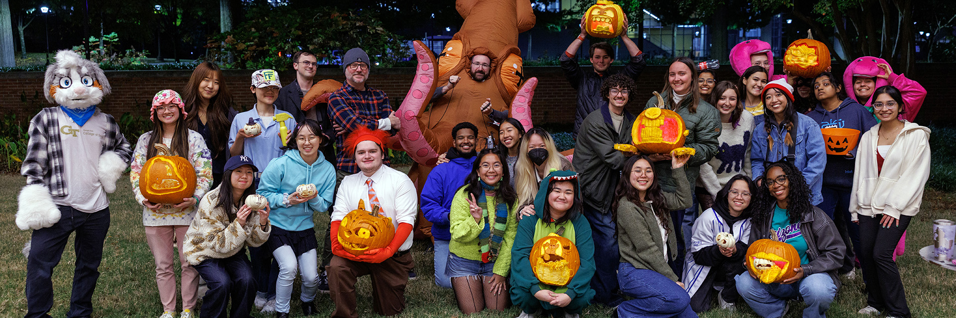 Photograph of students and faculty, some in Halloween costume, pose on the lawn of Georgia Tech's West Architecture building, holding carved pumpkins.