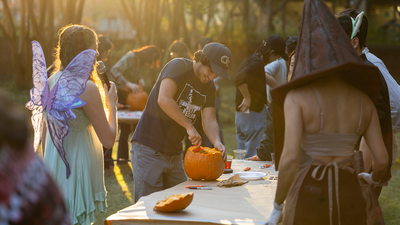 Student in fairy costume holds a mic to another student in a Tech hat as he guts a pumpkin