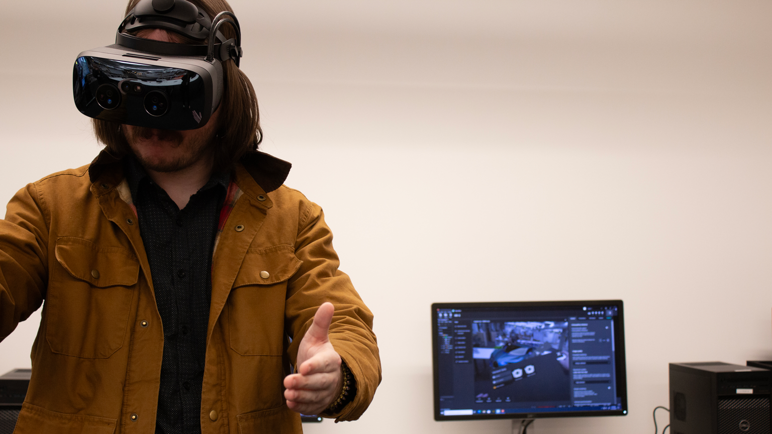 A man wearing a VR headset on the left side of the photo. To the right, a computer monitor displays what he sees.